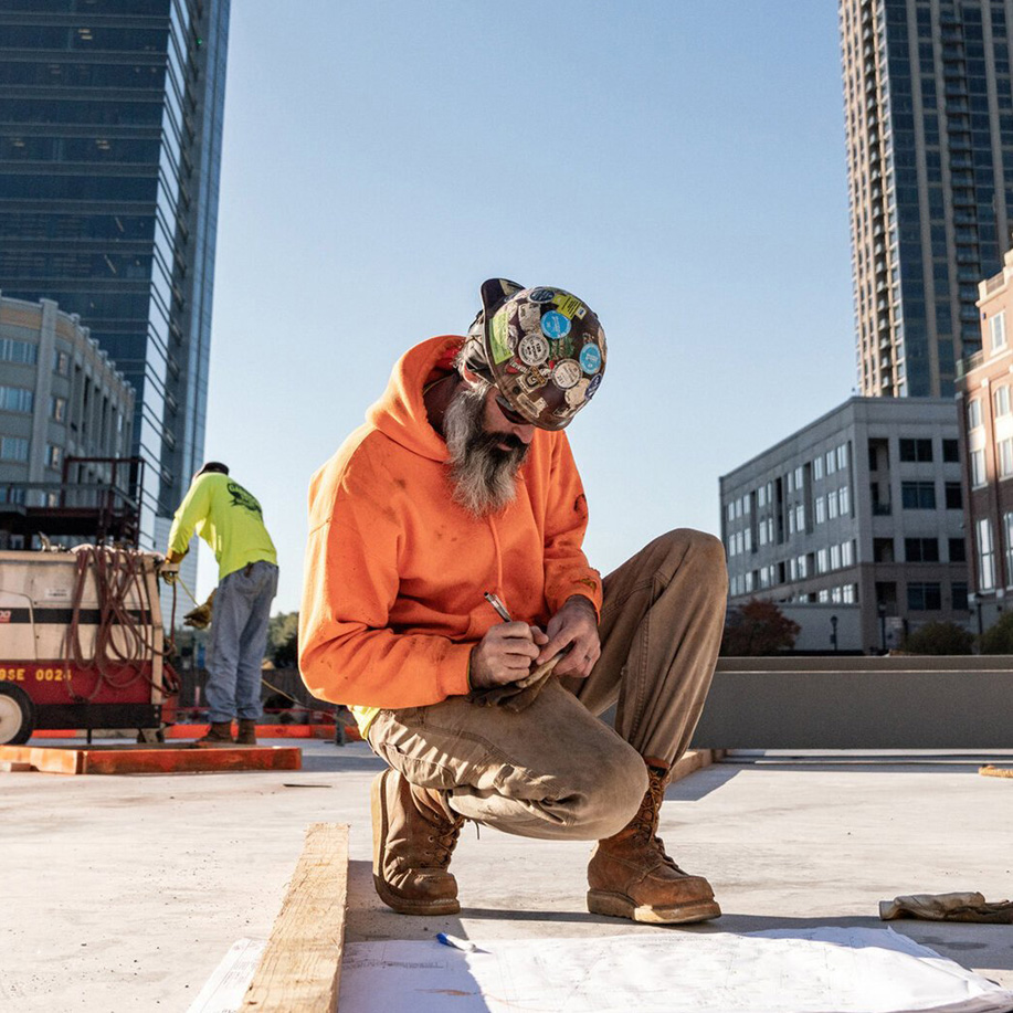 A tradesman in an orange hoodie crouched at work on a city rooftop